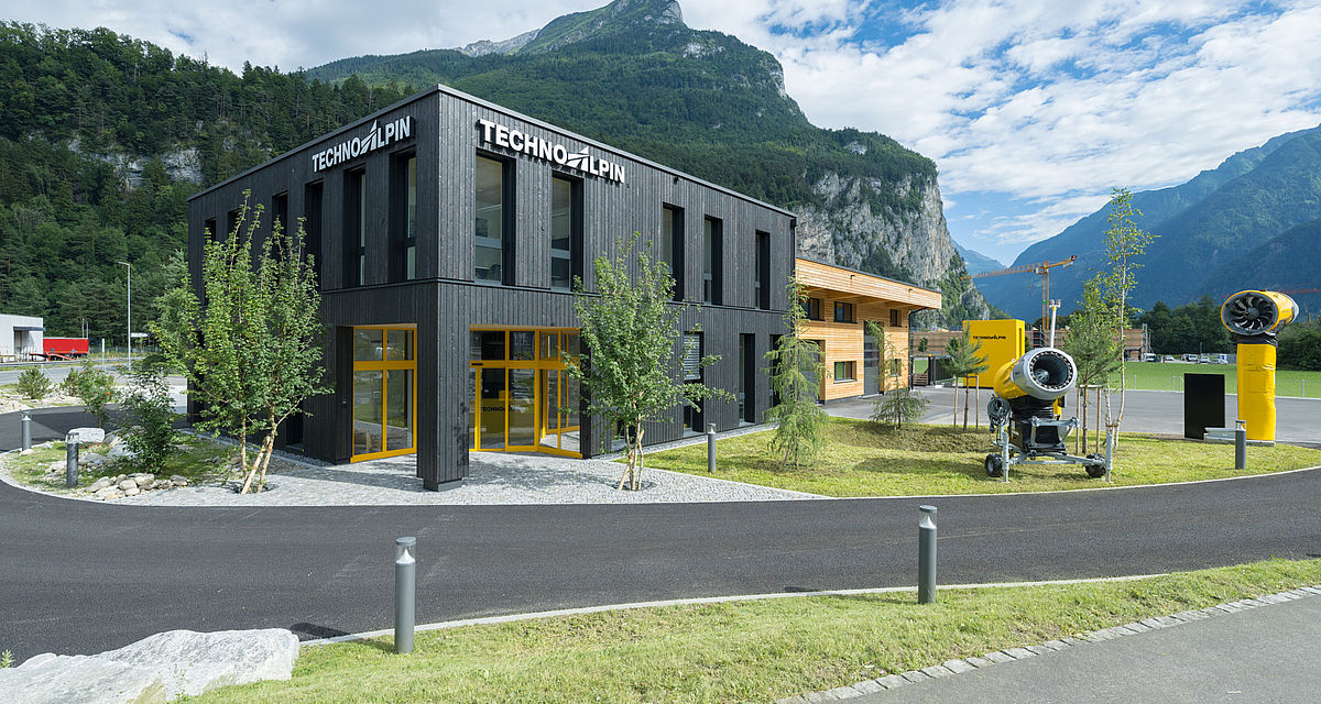 Modernes TechnoAlpin Firmengebäude mit Holzfassade und ausgestellten Schneeerzeugern vor Bergkulisse