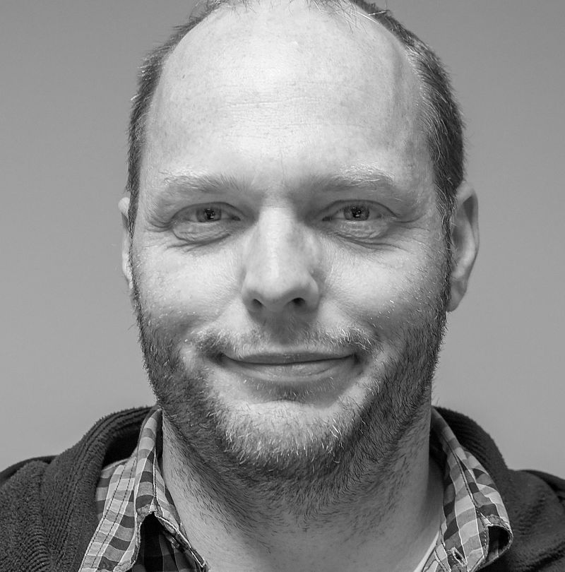 Marco Albiez, TechnoAlpin Mitarbeiter, Schwarz-Weiß-Portrait mit kurzem Haar, Bart und sanftem Lächeln in Fleecejacke über kariertem Hemd