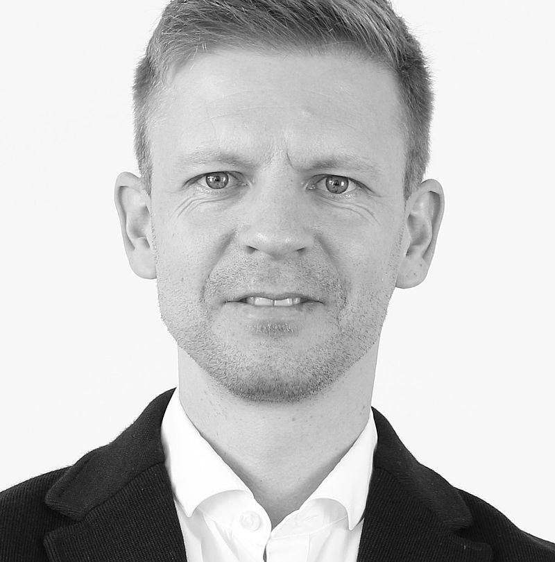 Matthias Illmer, TechnoAlpin Mitarbeiter, Schwarz-Weiß-Portrait mit kurzem blondem Haar, freundlichem Lächeln und dunklem Sakko über weißem Hemd