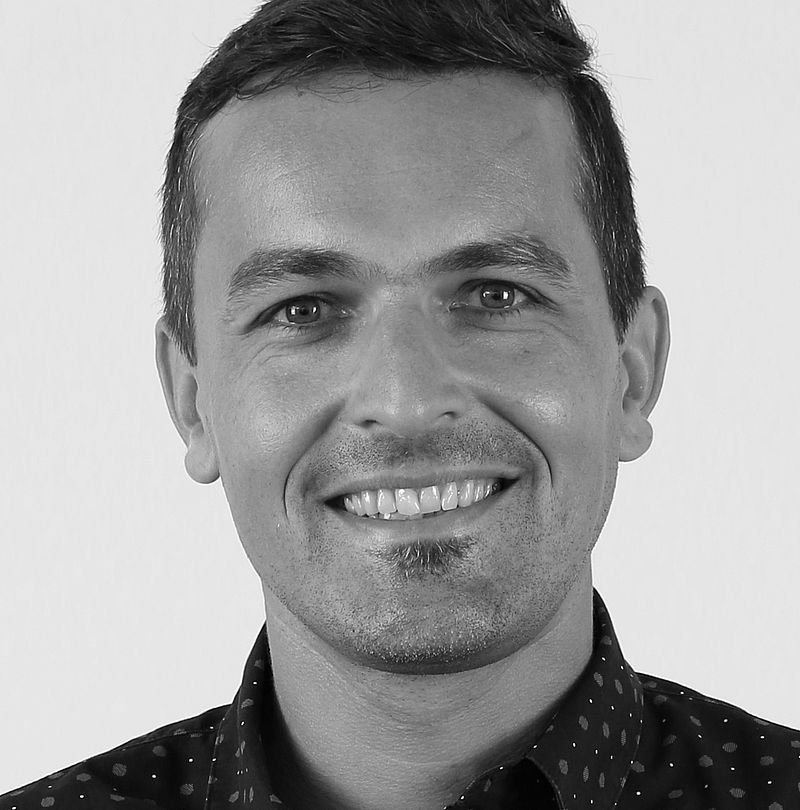 Alexander Aichorn, TechnoAlpin Mitarbeiter, Schwarz-Weiß-Portrait mit breitem Lächeln und dunklem gemustertem Hemd vor hellem Studiohintergrund