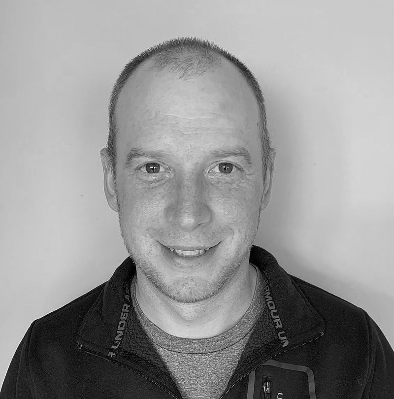 Aaron Madison, TechnoAlpin Mitarbeiter, Portrait mit sehr kurzem Haar, dezentralem Lächeln und dunkler Fleecejacke mit Under Armour Lanyard