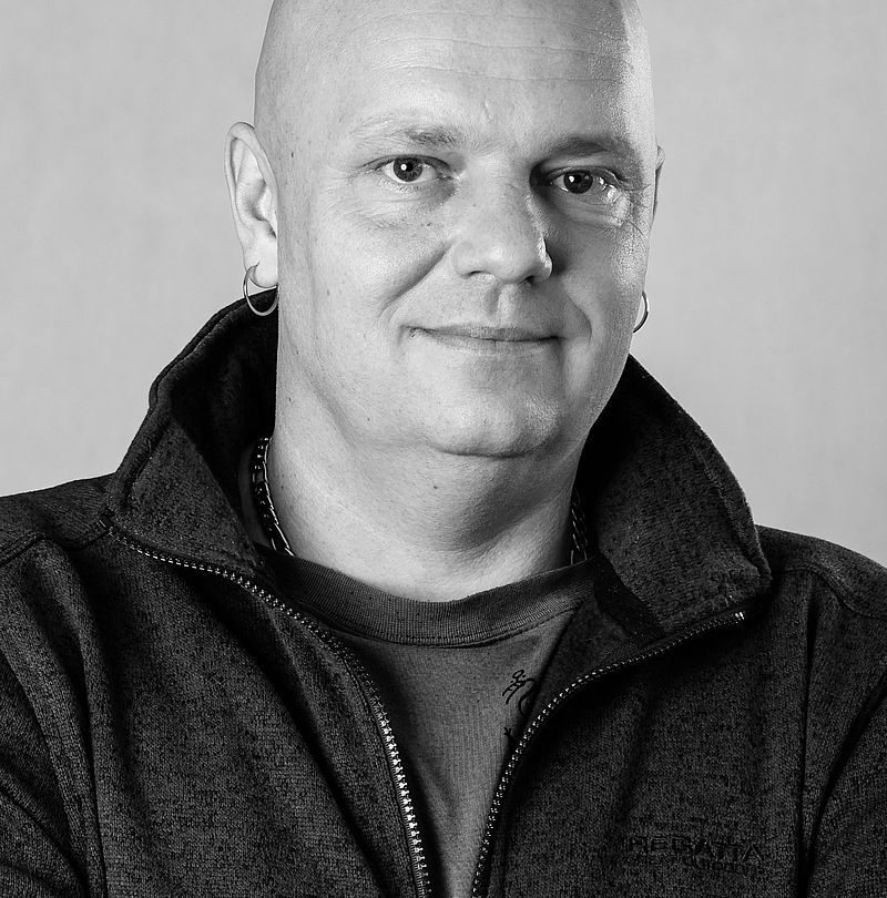 Martin Merka, TechnoAlpin Mitarbeiter, Schwarz-Weiß-Portrait mit Glatze, Ohrringe und dezentralem Lächeln in dunkler Fleece-Reißverschlussjacke