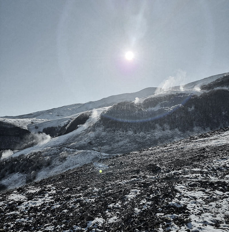Sonne mit Halo-Effekt über verschneitem Berghang mit Schneeerzeugern im Einsatz