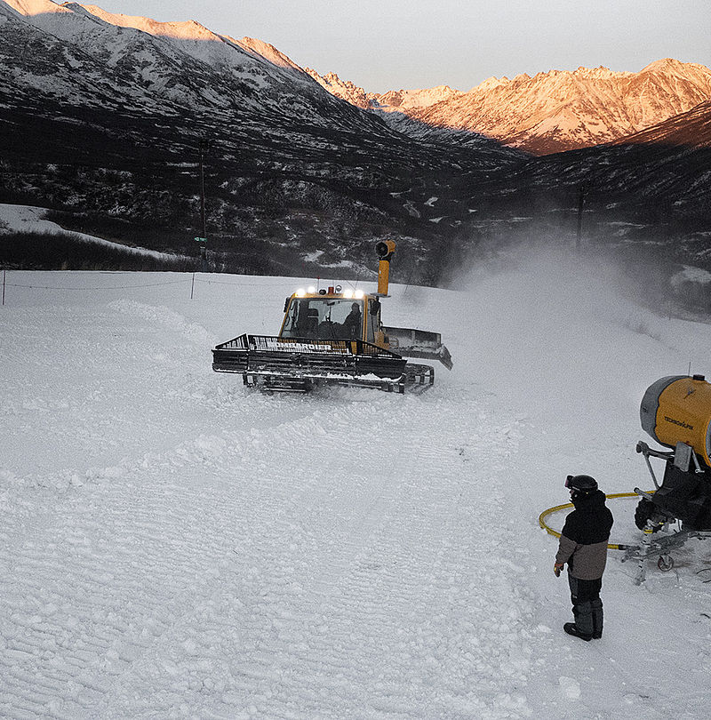 Piste groomer with mounted snow gun in action at dusk in mountain scenery