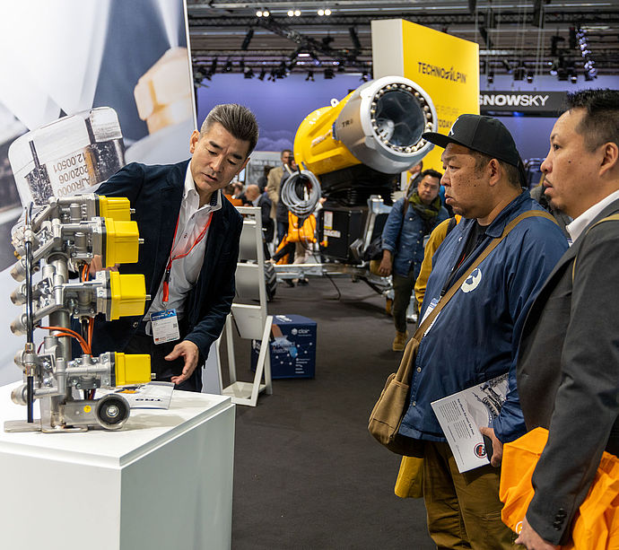 TechnoAlpin Mitarbeiter präsentiert Ventil-/Düsenbaugruppe asiatischen Besuchern auf der Interalpin, gelbe TR-Schneekanone im Hintergrund