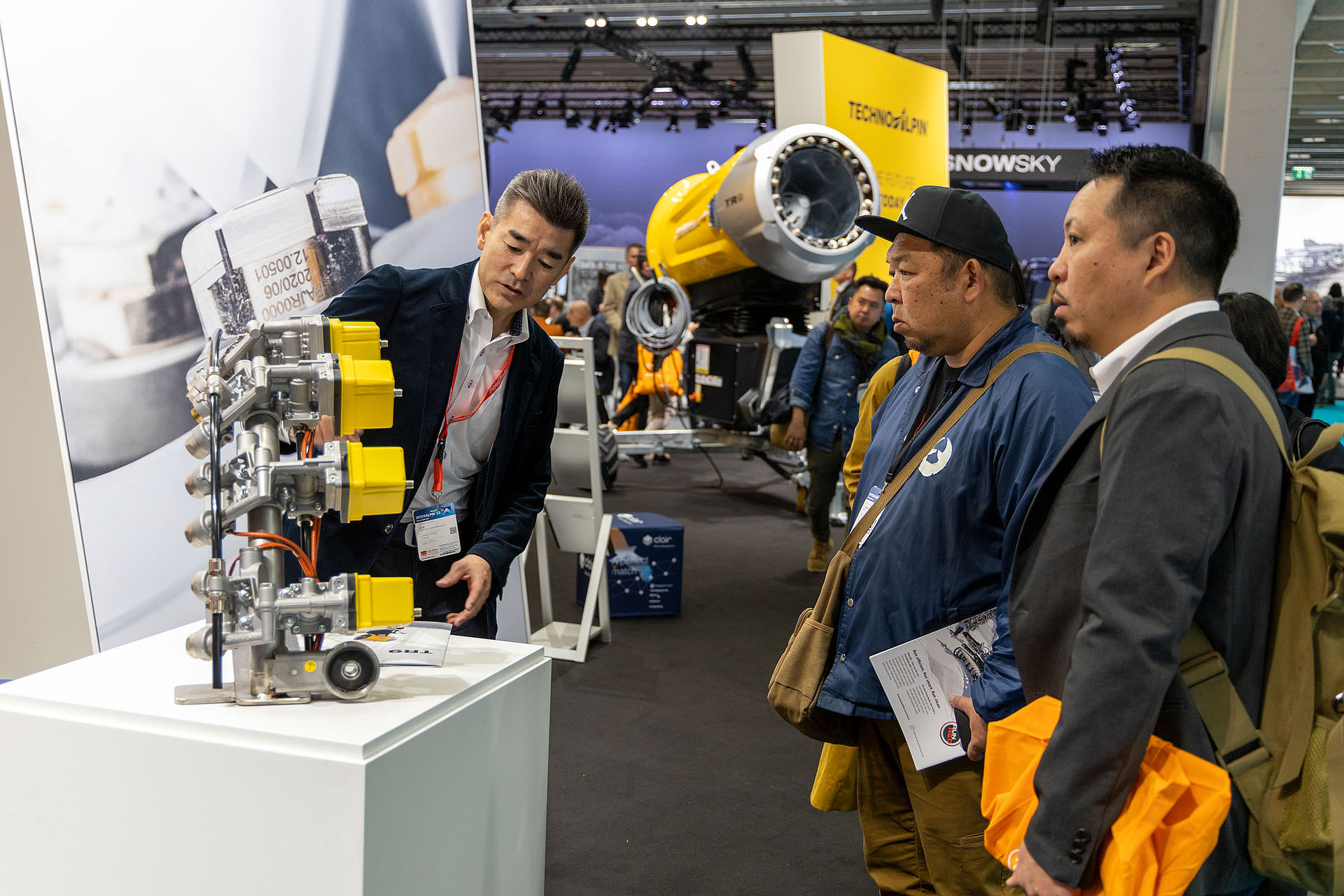TechnoAlpin Mitarbeiter präsentiert Ventil-/Düsenbaugruppe asiatischen Besuchern auf der Interalpin, gelbe TR-Schneekanone im Hintergrund