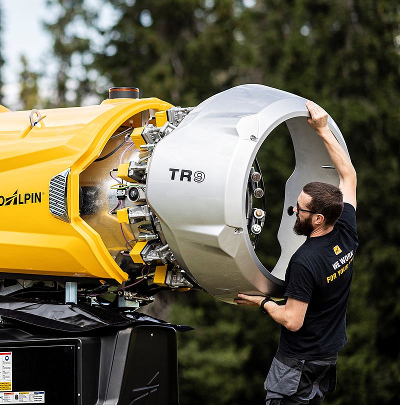 TechnoAlpin technician fitting silver TR9 nozzle ring onto yellow snow gun outdoors in forest setting