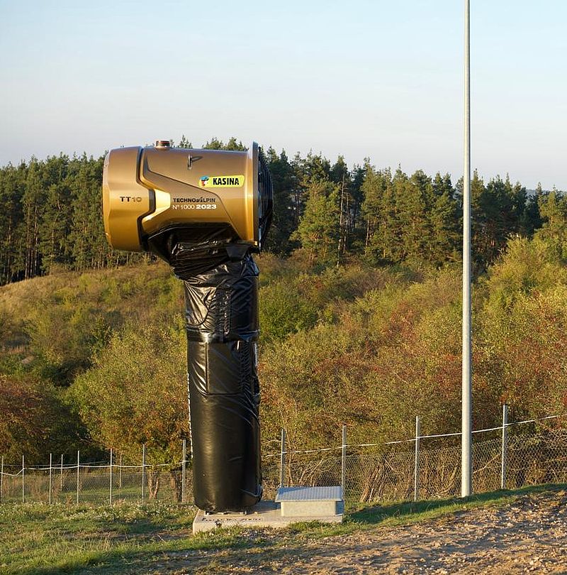 Goldene Jubiläums-Schneekanone TT10 mit Schutzhülle vor Waldlandschaft im Skigebiet
