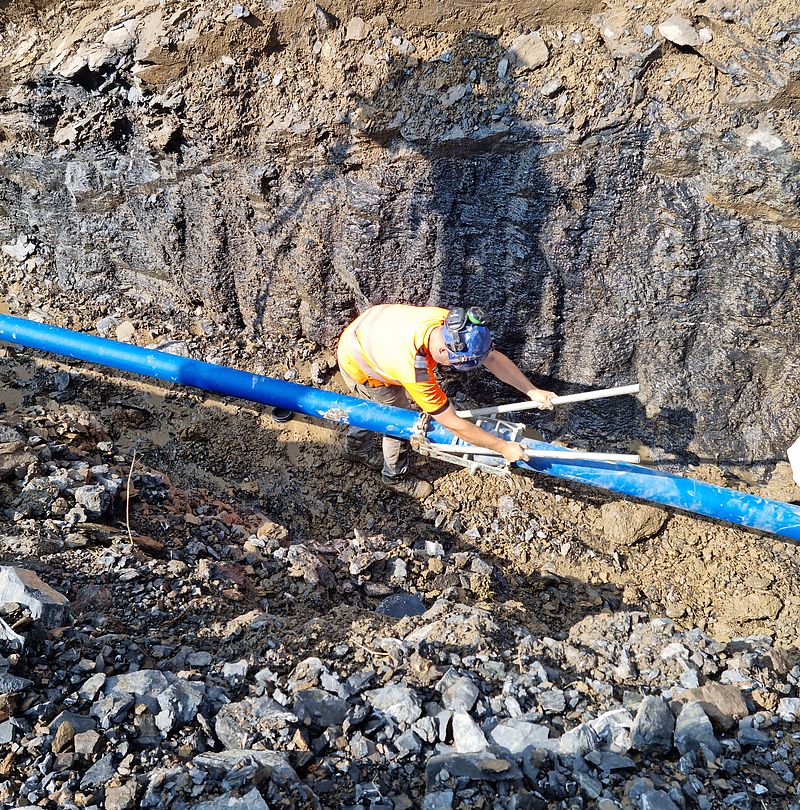 Bauarbeiter mit Helm und Warnweste arbeitet an blauen Rohrleitungen in steinigem Graben