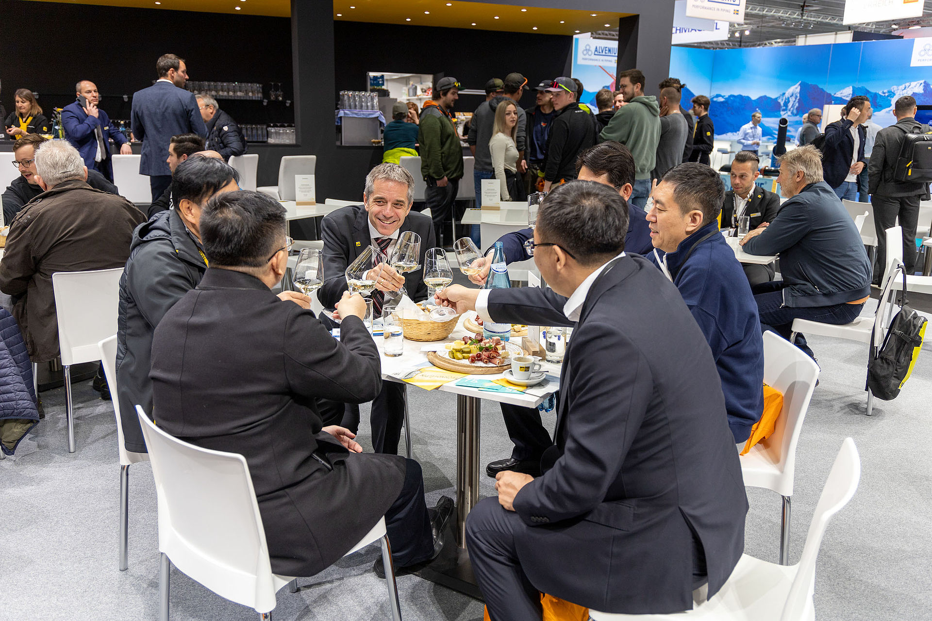 Besucher stoßen mit Weingläsern in der Hospitality-Lounge des TechnoAlpin Interalpin 2023 Standes an, Käseplatte auf dem Tisch