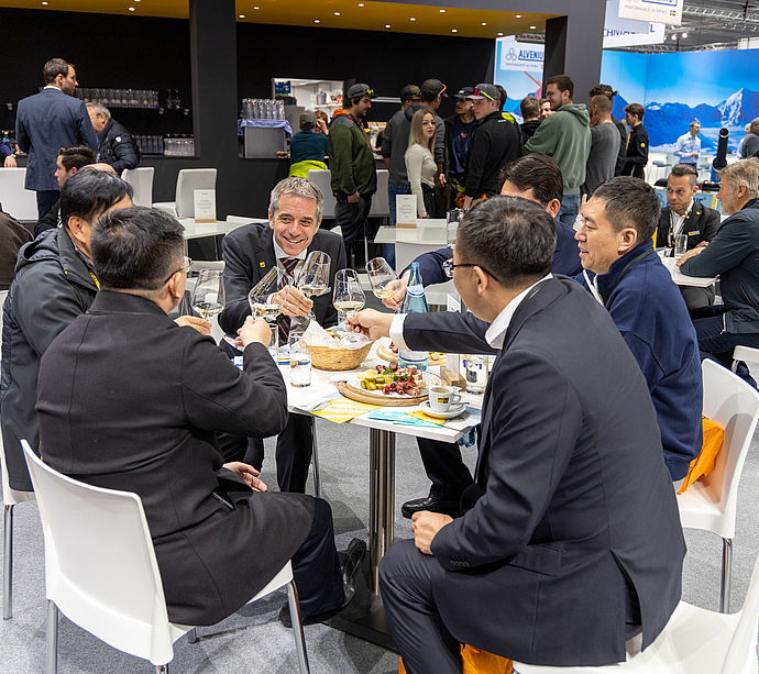 Besucher stoßen mit Weingläsern in der Hospitality-Lounge des TechnoAlpin Interalpin 2023 Standes an, Käseplatte auf dem Tisch