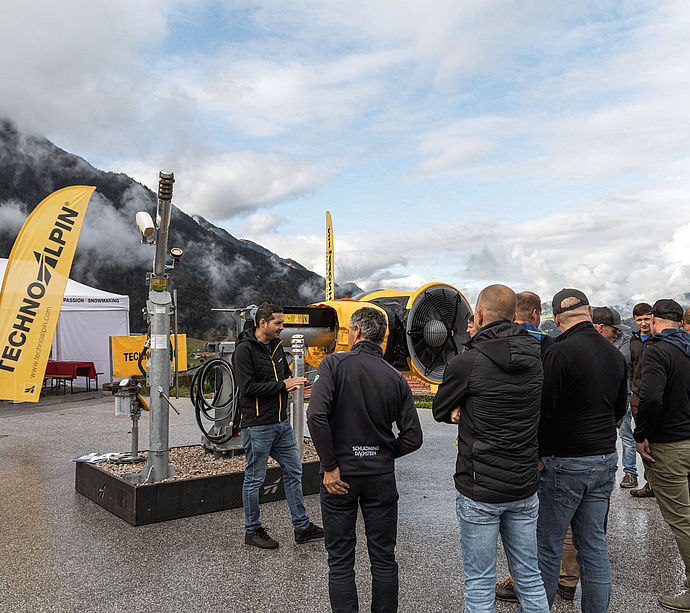 Roadshow TechnoAlpin all'aperto con gruppo visitatori davanti a innevatore TT10 e panorama alpino