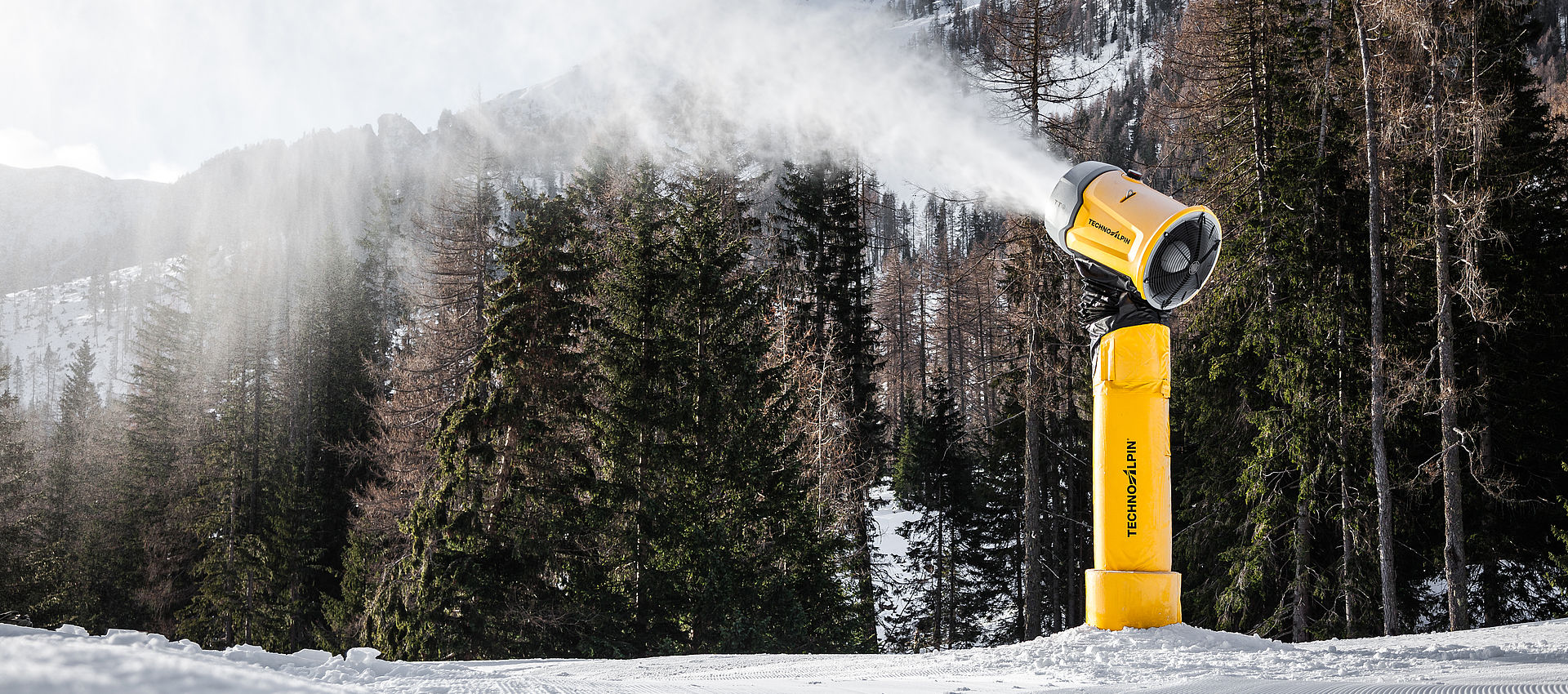 Gelbe Schneilanze TT9 erzeugt Schnee auf frisch präparierter Piste vor Bergwald und Alpengipfeln