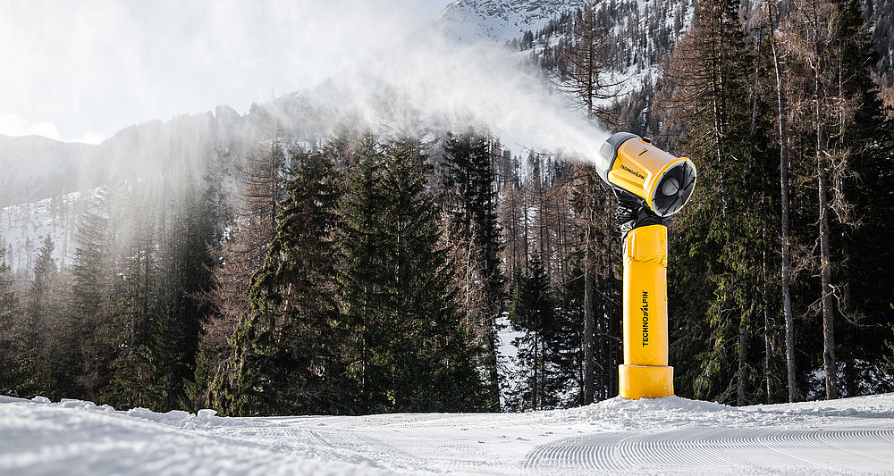 Gelbe Schneilanze TT9 erzeugt Schnee auf frisch präparierter Piste vor Bergwald und Alpengipfeln