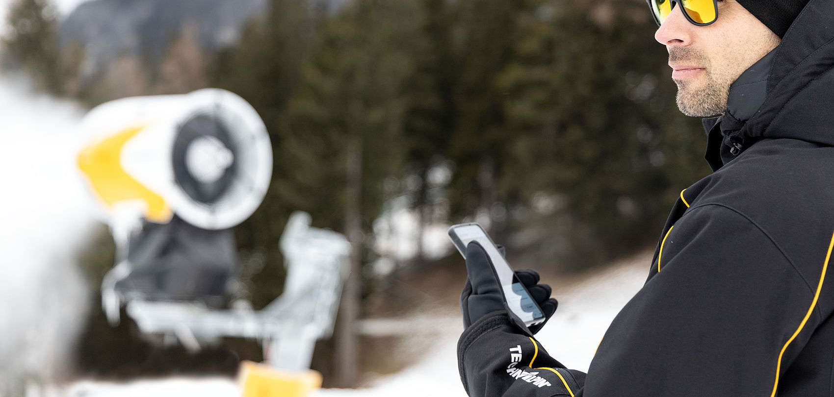 TechnoAlpin employee with sunglasses and smartphone in front of snow gun in snowy forest