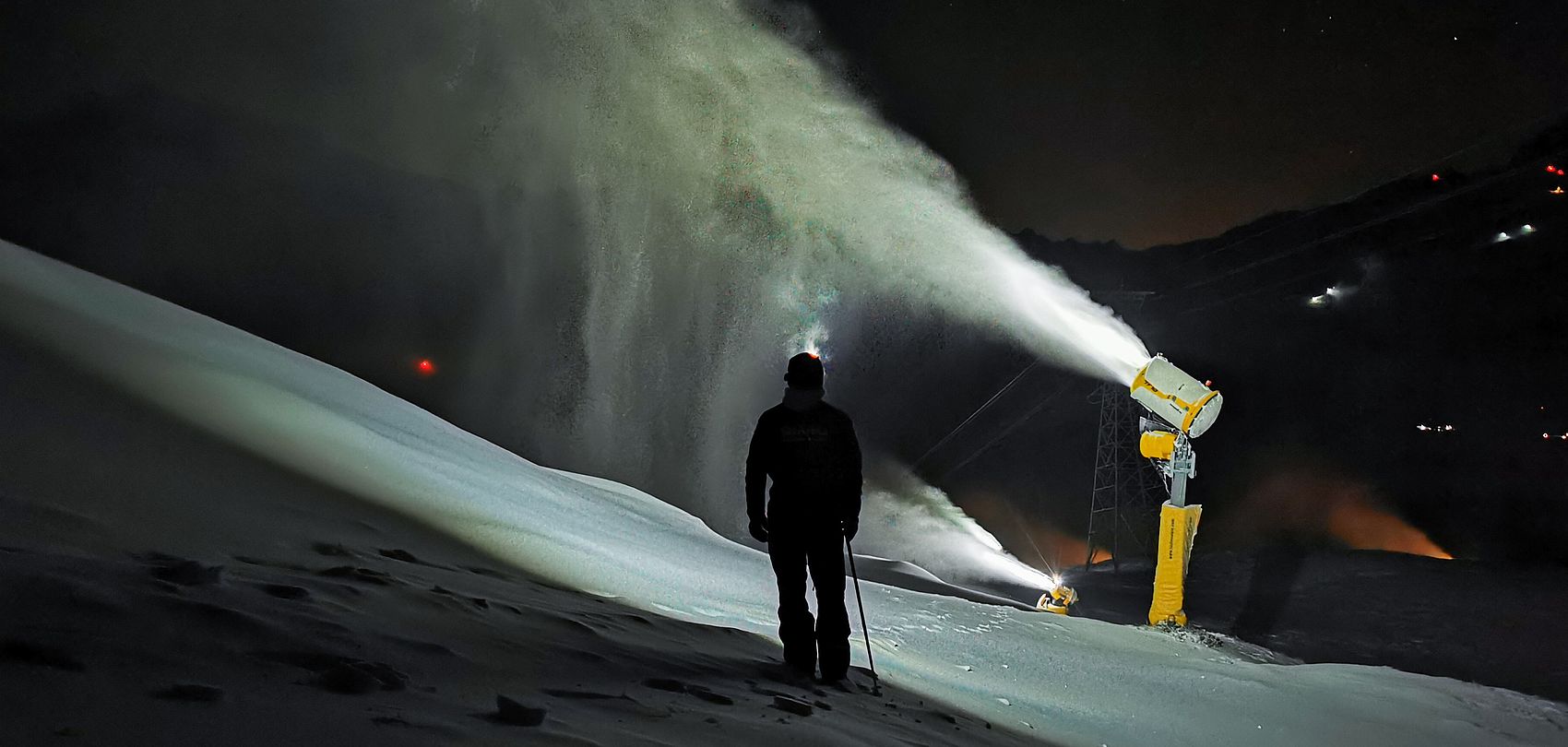 Silhouette of man in front of TechnoAlpin snow generator during nighttime snowmaking.