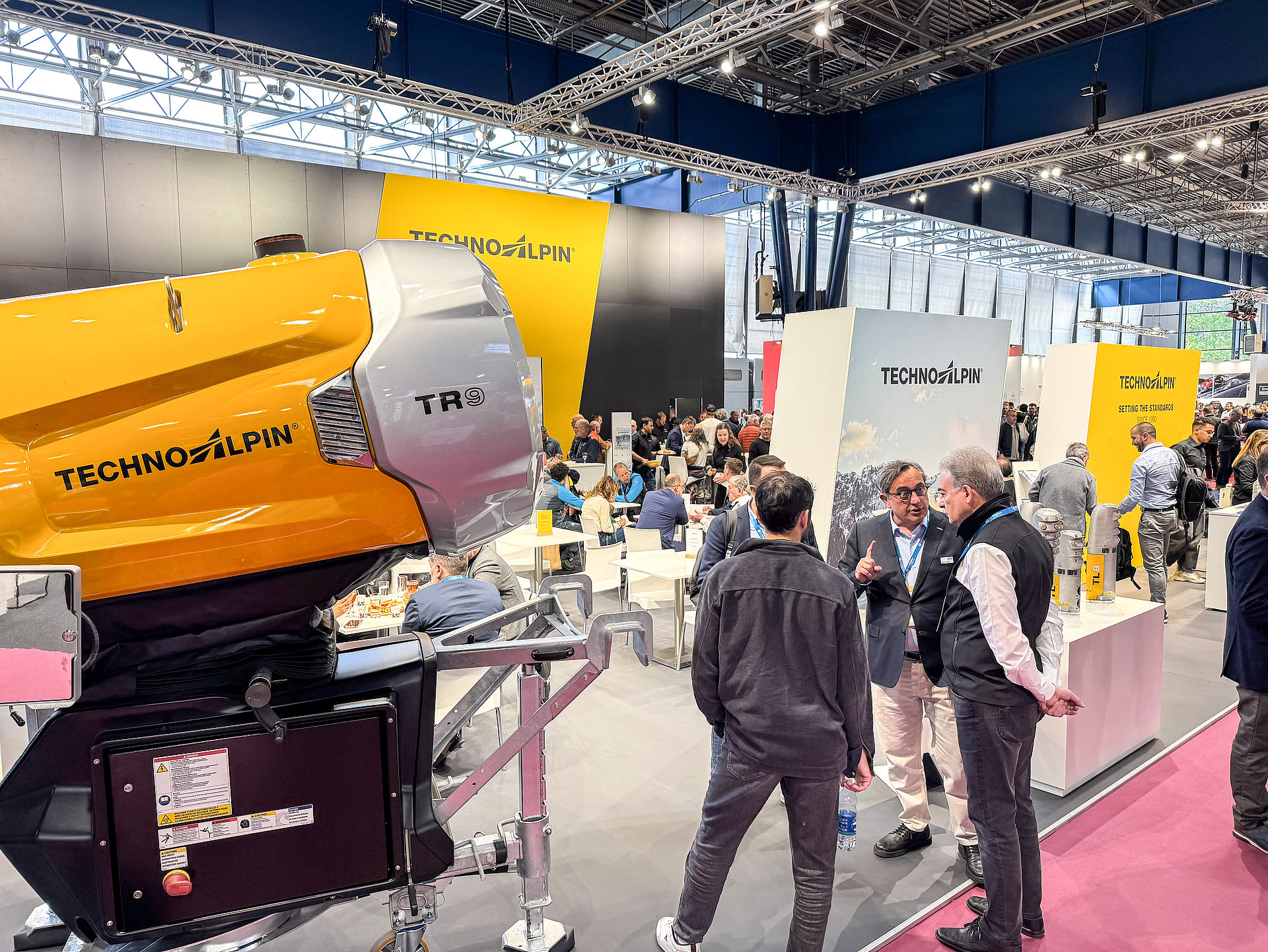 Gelber TR9 Propellerschneeerzeuger im Vordergrund, Berater im Gespräch am Messestand
