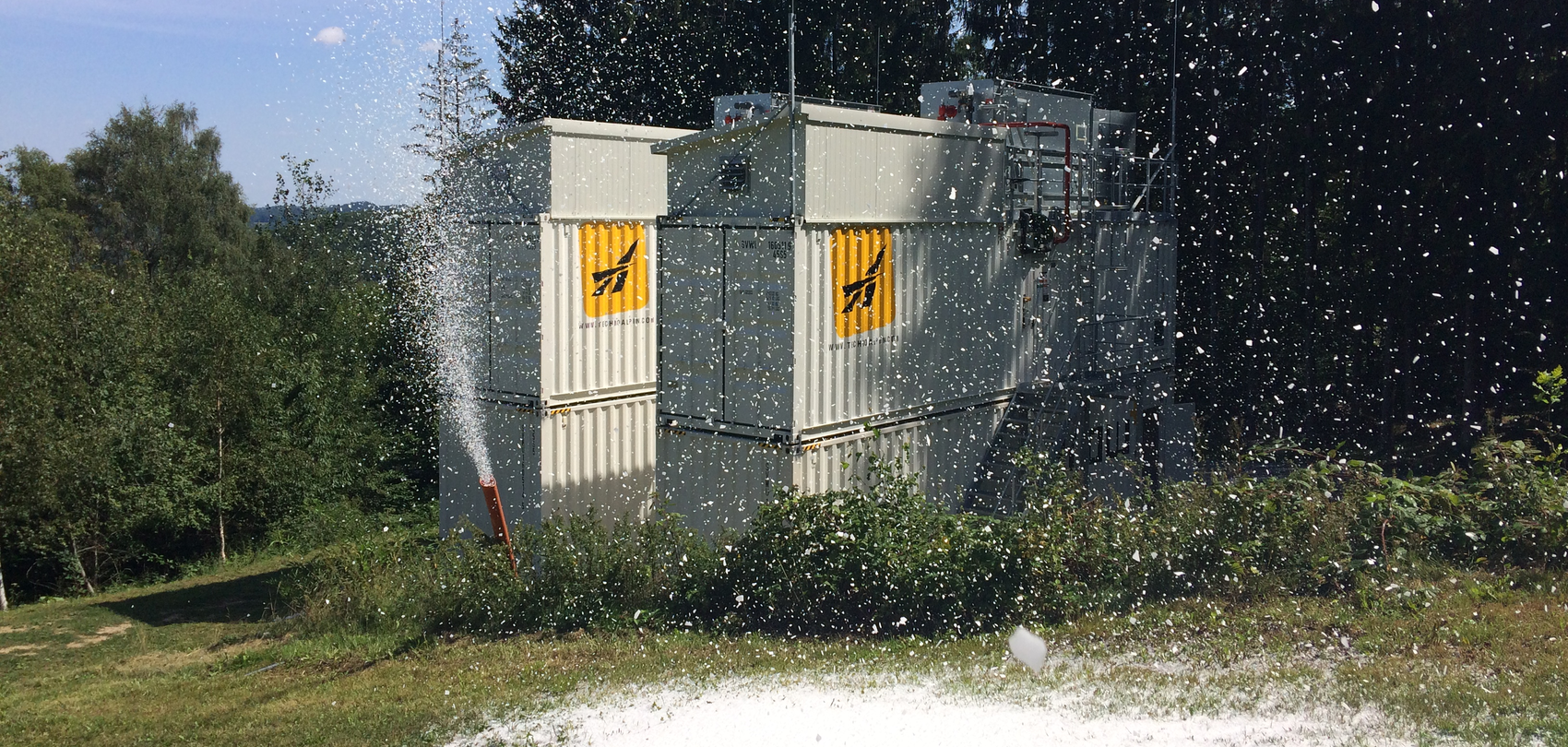 TechnoAlpin Indoor Snowmaker im Testbetrieb, Beschneiungsanlage produziert Schnee im Sommer auf grüner Wiese neben Containern im Wald