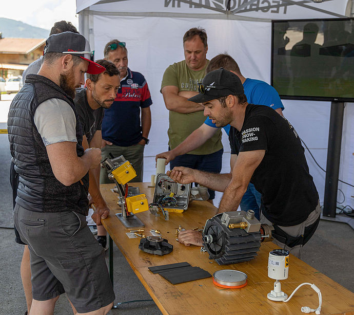 TechnoAlpin technician showing participants open control unit and components under pavilion.