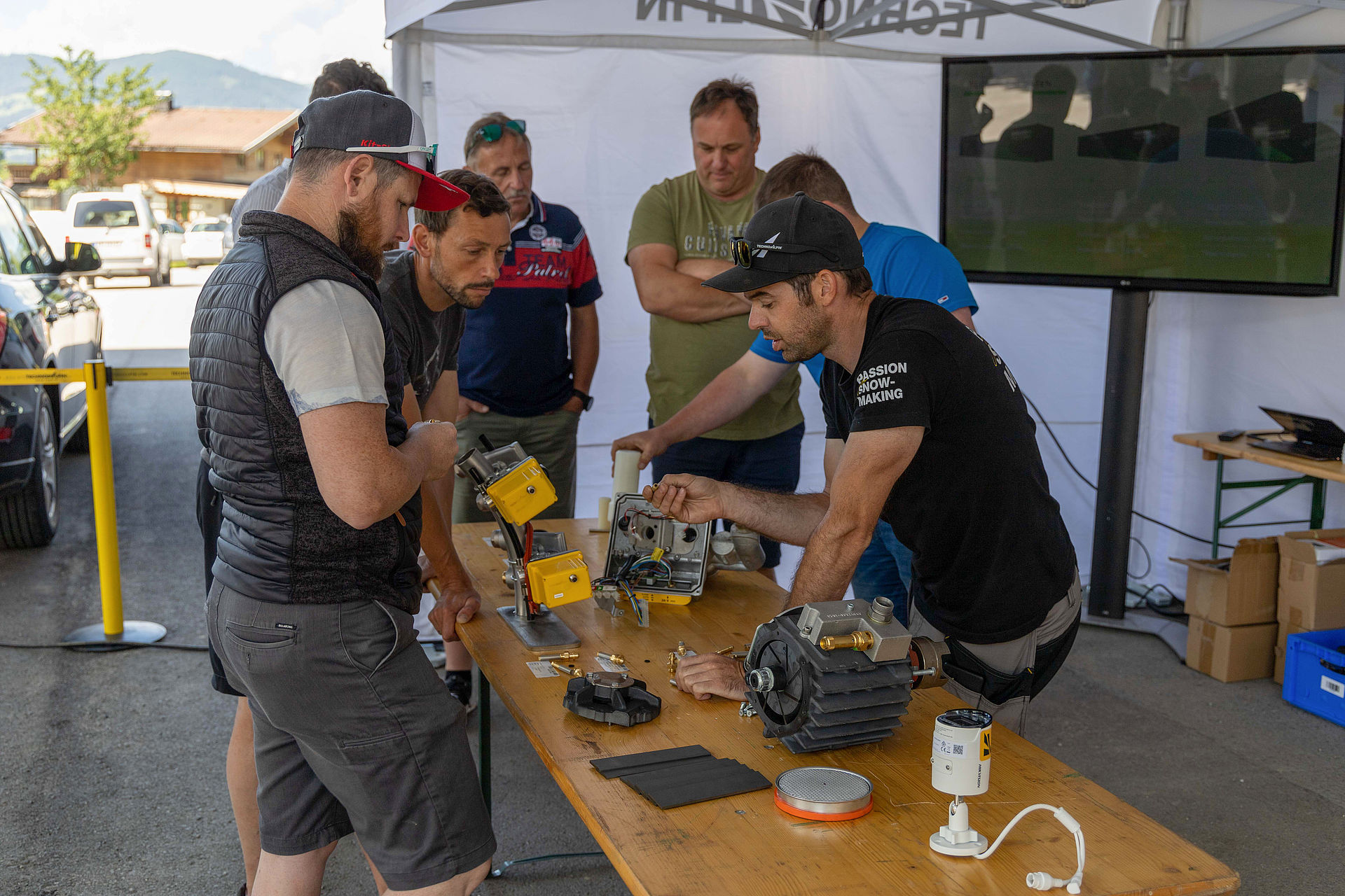 TechnoAlpin technician showing participants open control unit and components under pavilion.
