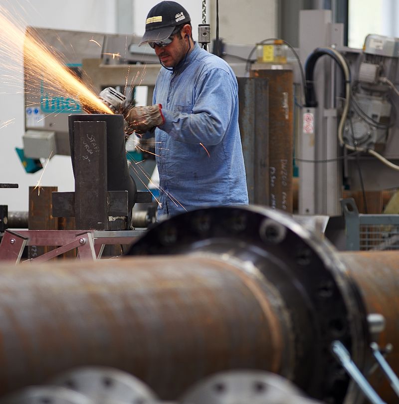 Worker with hat and safety goggles grinding steel component with sparks flying in workshop.