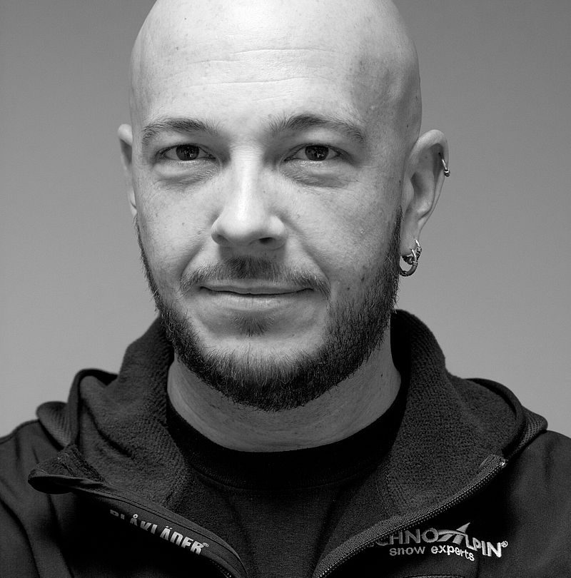 Ralf Gisler, TechnoAlpin Mitarbeiter, Schwarz-Weiß-Portrait mit Glatze, Bart und scherzhaftem Grinsen in TechnoAlpin Markenjacke