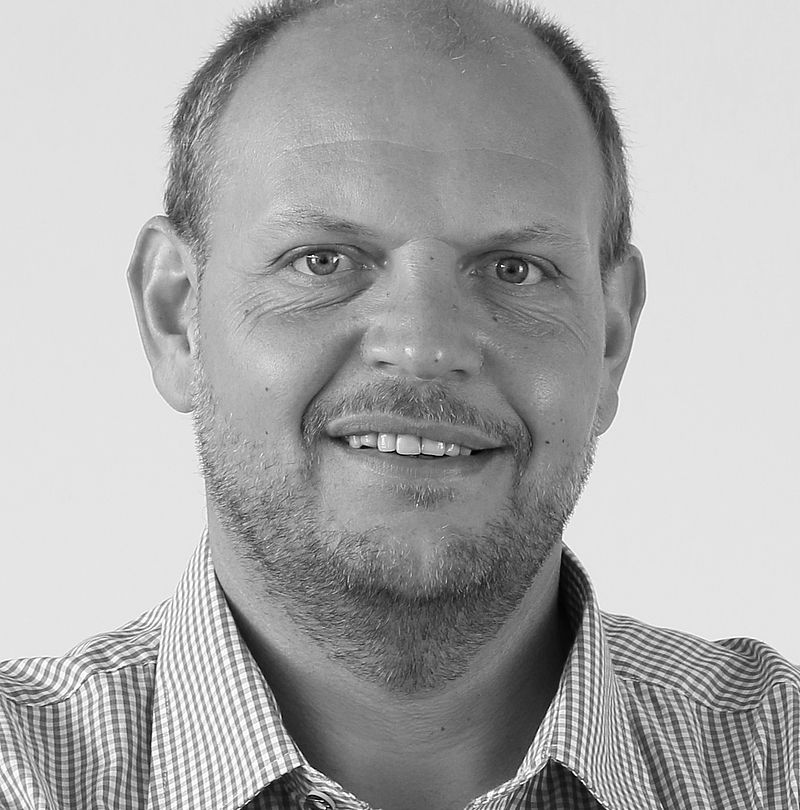 Albert Gomig, TechnoAlpin Mitarbeiter, Schwarz-Weiß-Portrait mit kurzem Haar, Dreitagebart und warmem Lächeln in kariertem Hemd