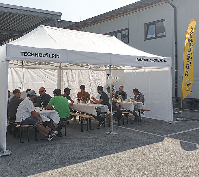 Visitatori del roadshow TechnoAlpin sotto tenda bianca a panche con beach flag giallo