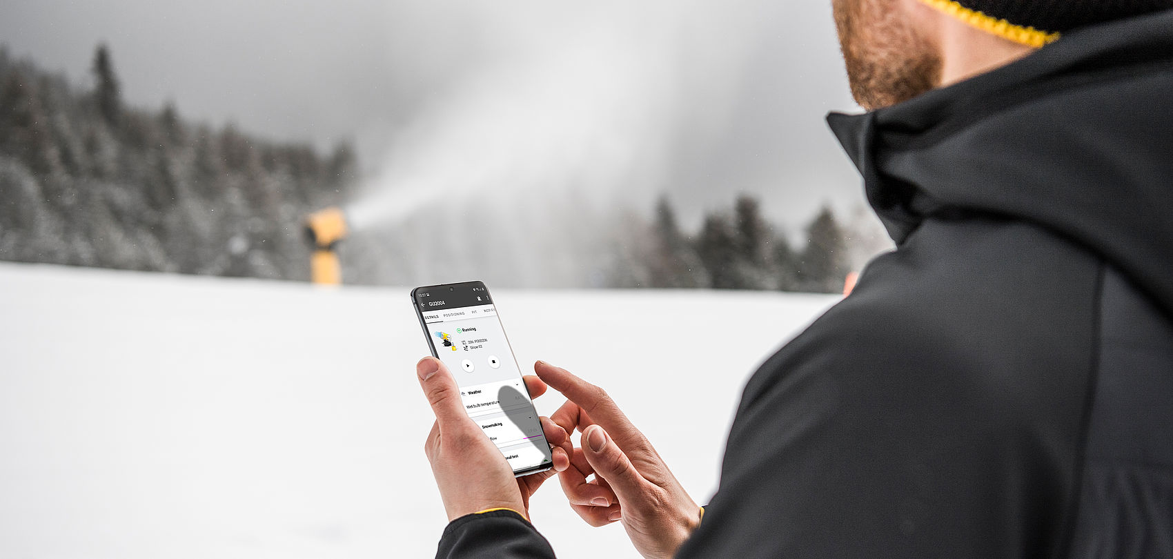 Mann mit Mütze und TechnoAlpin-Jacke bedient Smartphone-App vor schneeerzeugender Propellermaschine im Skigebiet