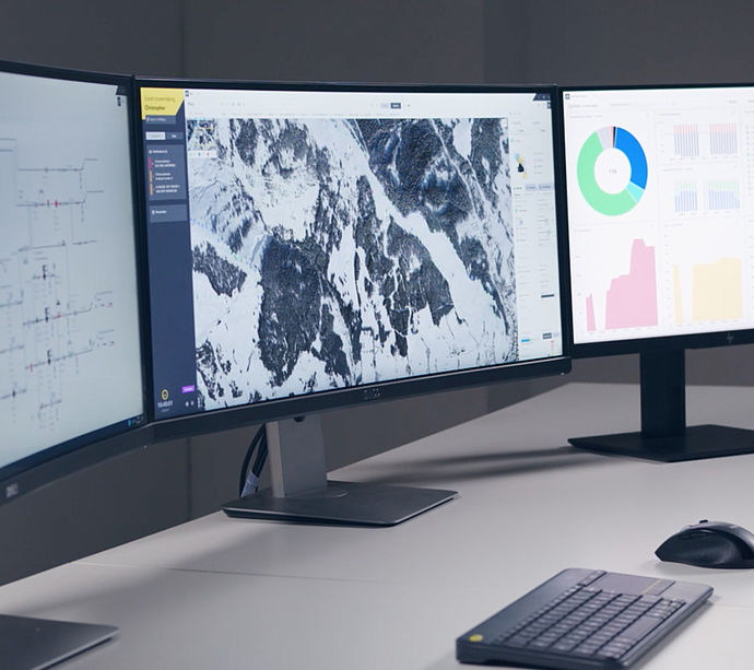 TechnoAlpin ATASSplus Schneeanlagensteuerung auf Drei-Monitor-Workstation mit Beschneiungsnetz-Schema, Skigebiets-Luftbild und Analyse-Dashboards