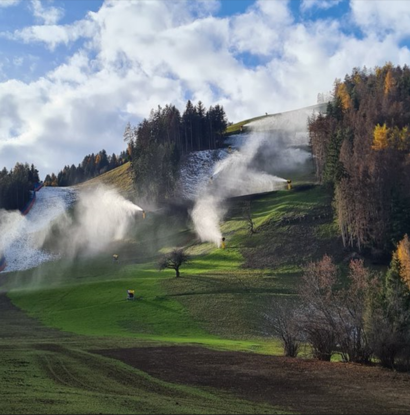 Schneeerzeuger im Einsatz auf grüner Skipiste am Berghang mit Herbstwald unter blauem Himmel mit Wolken