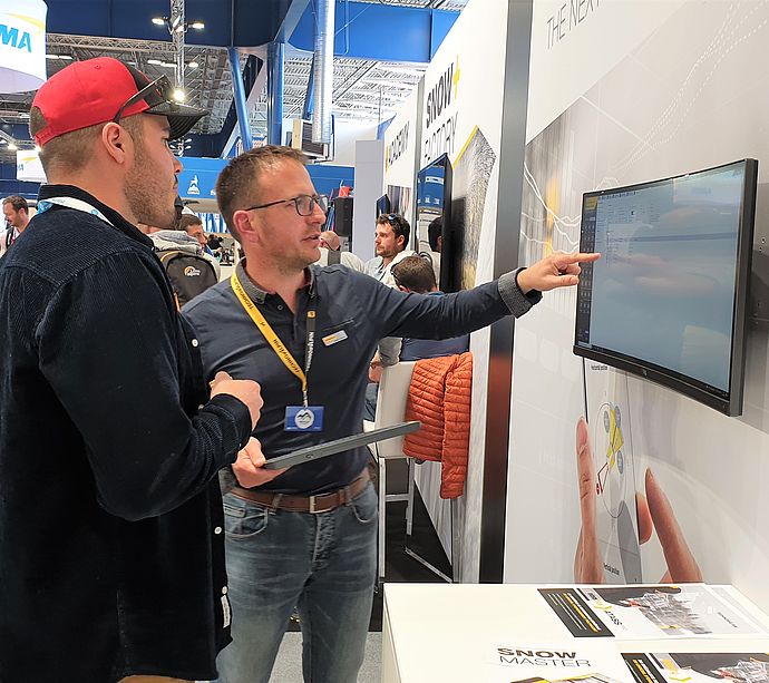 TechnoAlpin employee explaining software on touchscreen to visitor at trade show stand.