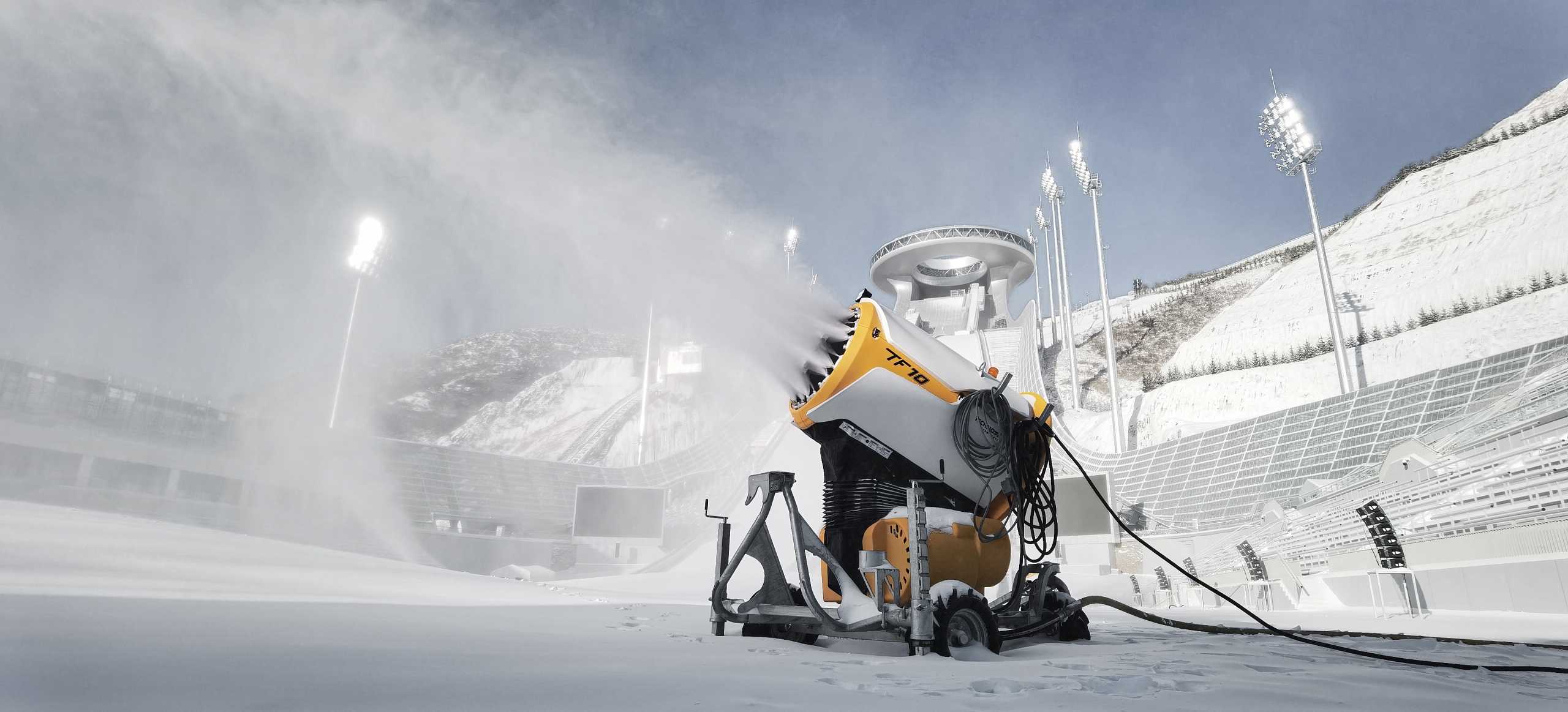 TechnoAlpin TF10 Propellermaschine erzeugt Schnee in Olympia-Skisprungstadion mit beleuchteten Flutlichtmasten